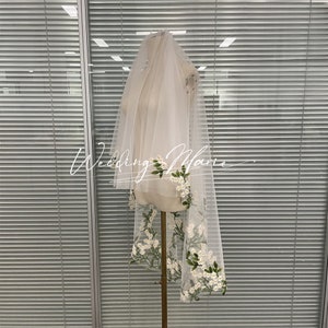 Puede incluir: Un velo de novia blanco transparente con delicado bordado floral. El velo presenta flores blancas en forma de margarita y hojas verdes a lo largo del borde inferior. El texto "Wedding Marie" está escrito en cursiva.