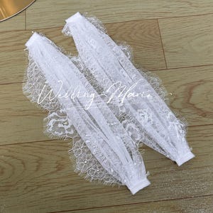 Op de afbeelding: Twee witte kanten bruidsmanchetten met delicate bloempatronen en een geschulpte rand. De tekst "Wedding Marie" is zichtbaar op de manchetten. De manchetten zijn gemaakt van een zachte, transparante stof.
