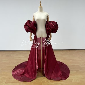 Puede incluir: Un vestido burdeos con una larga cola y mangas abullonadas. El vestido se exhibe en un maniqu&iacute; con un soporte de madera. El texto "Wedding Marie" es visible en la parte delantera del vestido.