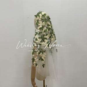 Op de afbeelding: Een witte bruidssluier met bloemenborduurwerk. De sluier is versierd met delicate witte bloemen en groene bladeren, wat een romantische en elegante uitstraling creëert. De tekst "Wedding Marie" staat op de sluier.