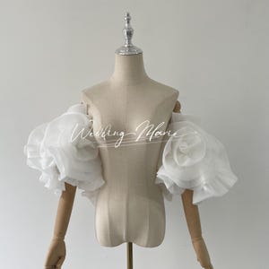 Può includere: Maniche da sposa bianche, a spalle scoperte, con grandi accenti floreali a balze. Le maniche sono realizzate in un tessuto trasparente e sono esposte su un manichino. Il testo "Wedding Marie" è scritto sul davanti.