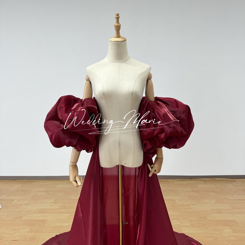 Puede incluir: Un vestido color burdeos con mangas abullonadas y voluminosas se exhibe en un maniqu&iacute;. El vestido presenta una falda larga y fluida y una superposici&oacute;n transparente. El texto "Wedding Marie" est&aacute; escrito en la parte delantera del vestido en cursiva.