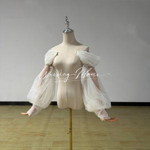 May include: A white, off-the-shoulder bridal top with sheer, puffy sleeves. The top is displayed on a mannequin. The text "Wedding Marie" is written across the front of the top in a cursive font.