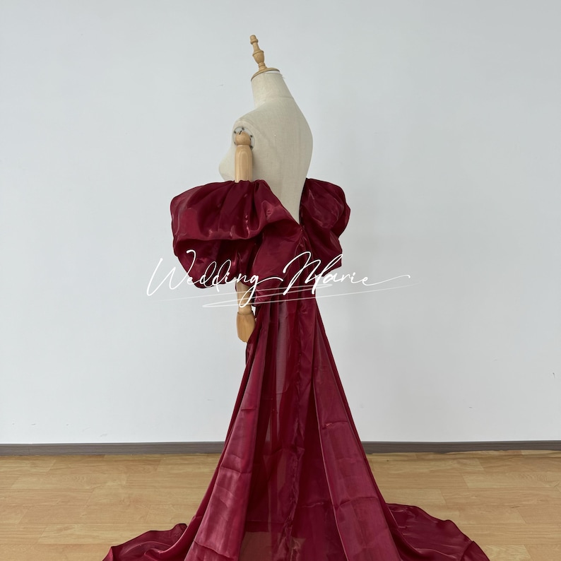 Puede incluir: Un vestido formal de color burdeos con un detalle de hombro dram&aacute;tico y de gran tama&ntilde;o. El vestido presenta una falda fluida y se exhibe en un maniqu&iacute;. El texto "Wedding Marie" est&aacute; escrito en el vestido.