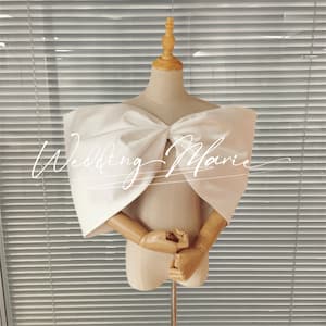 May include: A white satin bridal bolero with a large bow at the front. The bolero is on a dress form against a white blind background. The text "Wedding Marie" is written in white cursive on the bolero.