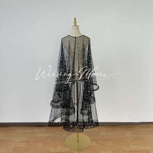 May include: A black tulle wedding cape with a scattered white star pattern. The cape is draped over a dress form and has a flowing, layered design.
