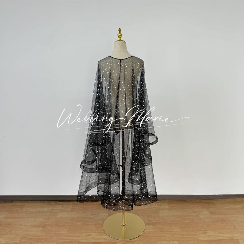 May include: A black tulle wedding cape with a scattered white star pattern. The cape is draped over a dress form and has a flowing, layered design.