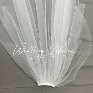 May include: A white tulle wedding veil with a single row of white buttons at the bottom. The veil is folded in half and the text "Wedding Marie" is written in cursive on the tulle.
