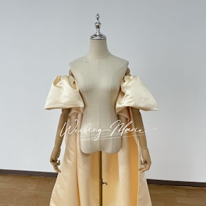 May include: A cream-colored satin dress with large, puffy sleeves and a long train. The dress is displayed on a mannequin.