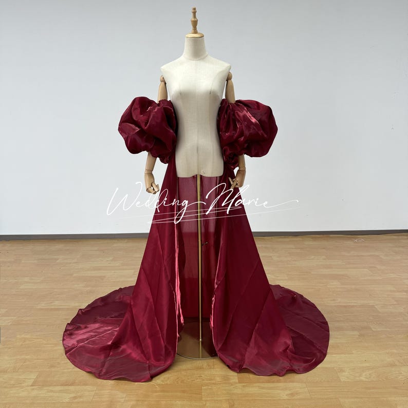 Puede incluir: Un vestido burdeos con una larga cola y mangas abullonadas. El vestido se exhibe en un maniqu&iacute; con un soporte de madera. El texto "Wedding Marie" es visible en la parte delantera del vestido.