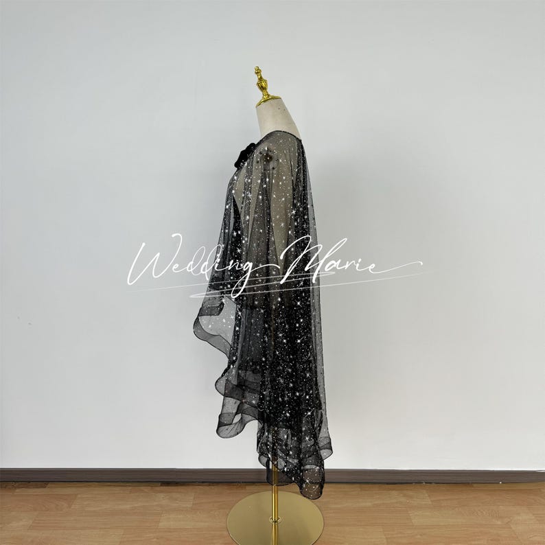 May include: Black sheer cape with a sparkly finish. The cape is draped over a mannequin and has a ruffled hem.