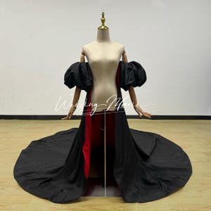 May include: A black and red formal gown with a long train, displayed on a mannequin. The gown features a red interior and black exterior, with large, puffy black sleeves. The mannequin is set against a plain white background.