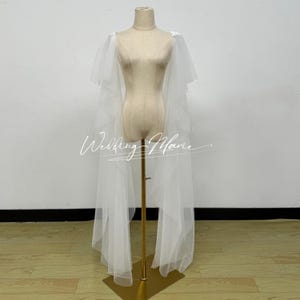 May include: A white tulle bridal cape with short sleeves and a long train, displayed on a mannequin. The cape is sheer and flowing, with the text "Wedding Marie" in a script font.