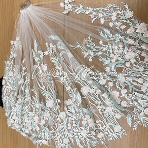 May include: A white bridal veil with delicate floral embroidery in shades of green and white. The veil is made of soft tulle and features a delicate floral pattern.