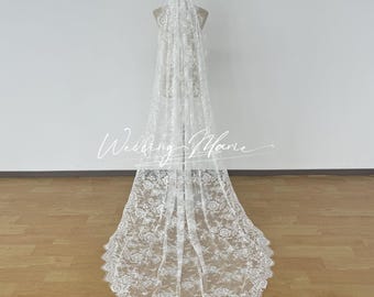 May include: A long, white lace wedding veil with a floral pattern and scalloped edge is displayed on a mannequin. The veil is sheer and delicate, with the text "Wedding Marie" in a script font.
