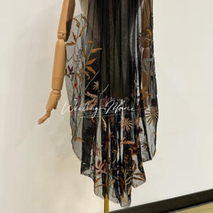 Op de afbeelding: Zwarte doorschijnende stof met gouden, bruine en rode bloemenborduurwerk. De stof is over een mannequin gedrapeerd, wat het gedetailleerde ontwerp en de textuur benadrukt. De tekst "Wedding Marie" is zichtbaar.