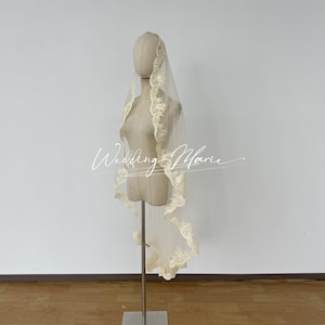 May include: A sheer, ivory-colored bridal veil with delicate lace trim, displayed on a mannequin. The veil is draped to showcase its length and design. The text "Wedding Marie" is visible.