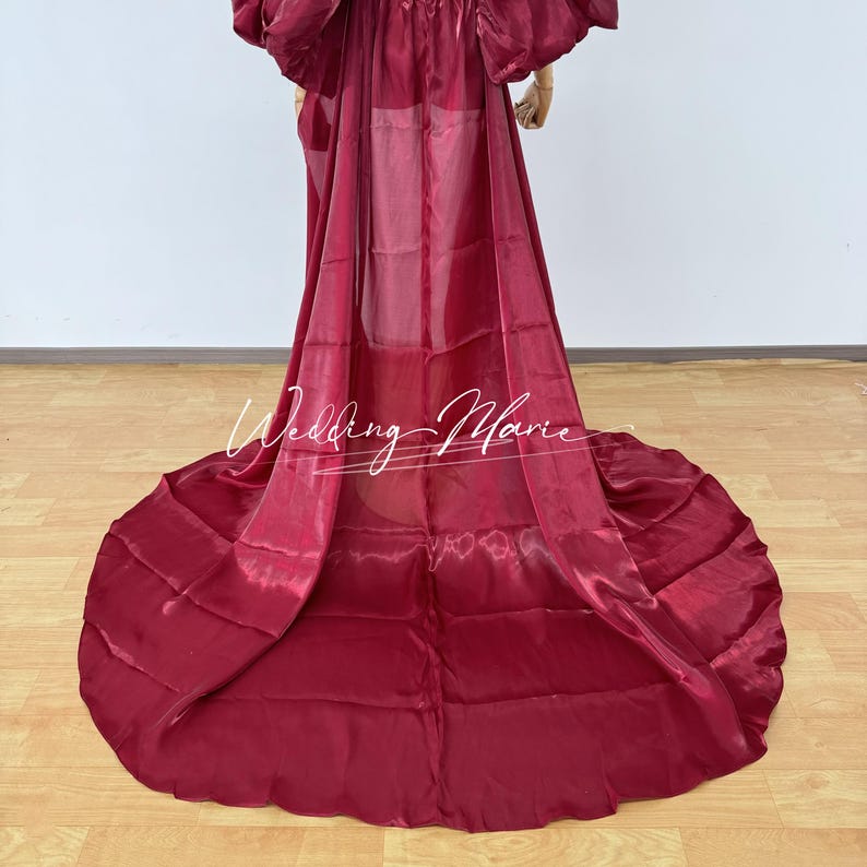 Puede incluir: Un vestido de color burdeos con una larga cola. La tela tiene brillo y la cola est&aacute; recogida en un patr&oacute;n circular. El texto "Wedding Marie" est&aacute; escrito en cursiva en la parte delantera del vestido.
