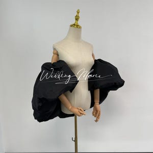May include: A black, voluminous sleeve garment displayed on a mannequin. The sleeves are gathered at the shoulder and flare out dramatically. The mannequin is beige, with a gold-colored head and wooden arms. The text "Wedding Marie" is written on the garment.