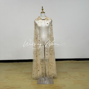 May include: A sheer, beige cape with star and floral embellishments. The cape is displayed on a mannequin and features a high collar and a flowing design. The cape is adorned with gold-coloured sequins and beads, creating a shimmering effect.