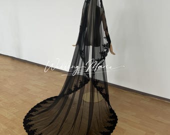 May include: A long, black bridal veil with intricate floral lace detailing along the edges. The veil is sheer, allowing a glimpse of the mannequin beneath. The train extends gracefully, creating a dramatic effect. The text "Wedding Marie" is visible.