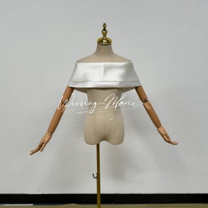 May include: A white satin off-the-shoulder top displayed on a mannequin. The top has a wide, folded neckline and is designed to be worn off the shoulders. The mannequin has wooden arms and a gold-colored stand. The text "Wedding Marie" is written across the front.