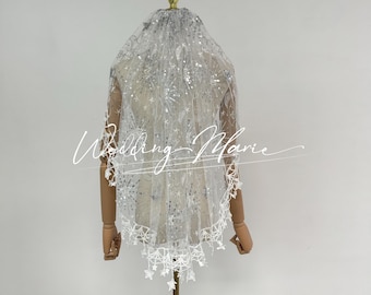 May include: A white bridal veil with a delicate lace trim and sparkling embellishments. The veil is displayed on a mannequin, showcasing its intricate design and flowing fabric. The text "Wedding Marie" is written on the veil.