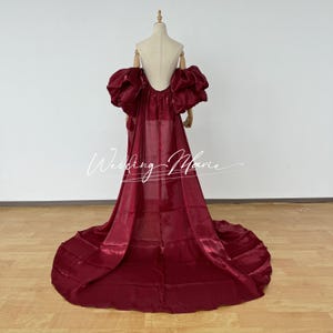 Puede incluir: Un vestido de noche burdeos con una larga cola y espalda descubierta se exhibe en un maniqu&iacute;. El vestido presenta mangas abullonadas grandes. El texto "Wedding Marie" est&aacute; escrito en cursiva.