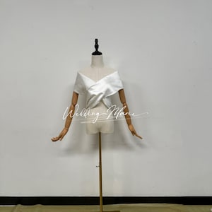 May include: A white off-the-shoulder top featuring a crossover wrap design across the chest, displayed on a mannequin with wooden arms. The garment has a smooth, solid finish and is positioned on a gold-coloured metal stand. The text Wedding Marie is visible.