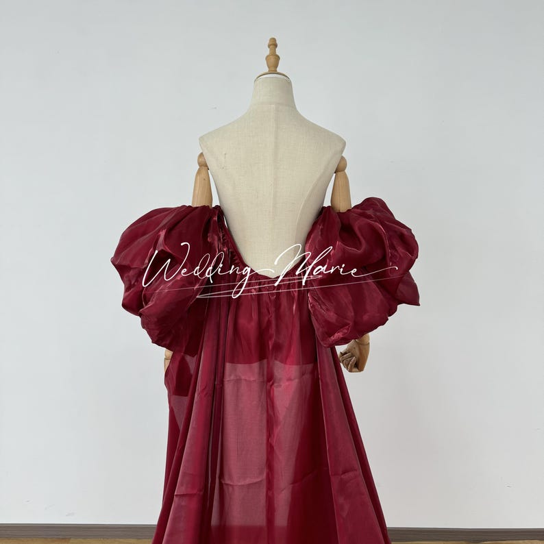 Puede incluir: Un vestido formal de color burdeos con mangas abullonadas y voluminosas. El vestido presenta una falda fluida y se exhibe en un maniqu&iacute;. El texto "Wedding Marie" est&aacute; escrito en cursiva en el vestido.