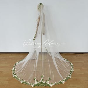 May include: A long, sheer white bridal veil with a floral trim of white flowers and green leaves. The veil is displayed on a mannequin and features the text "Wedding Marie".