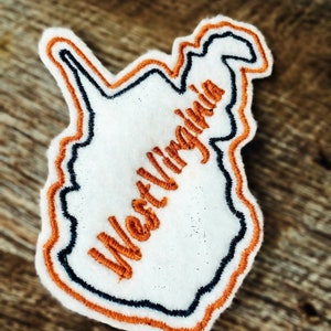 Puede incluir: Un parche de fieltro blanco con un contorno naranja y un contorno negro. El parche tiene la forma del estado de Virginia Occidental y tiene el texto "West Virginia" bordado en naranja.