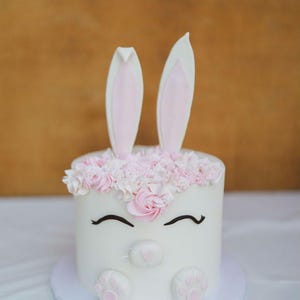 Puede incluir: Un pastel blanco decorado para parecerse a un conejito. El pastel presenta dos orejas verticales, huellas de patas rosas y un arreglo floral. La cara del conejito tiene pestañas negras curvas y una nariz rosa.