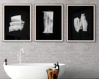 May include: Three black and white framed abstract art prints with white brushstrokes on a black background hang above a white bathtub and a wooden stool in a bathroom.
