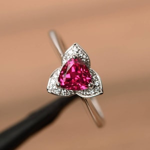 Puede incluir: Un anillo de plata con una piedra roja triangular rodeada de piedras blancas más pequeñas. El anillo tiene un diseño delicado con tres pétalos.