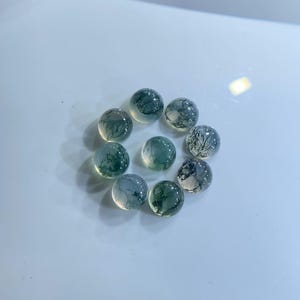 May include: Ten clear, round gemstones with green and white inclusions, arranged in a circle. The polished gemstones reflect light. Each stone is approximately 1 cm in diameter.