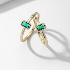 May include: Two gold rings with emerald green rectangular gemstones and small clear gemstones. The rings are intertwined.