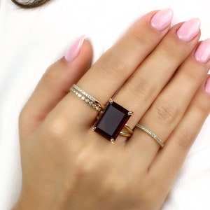 May include: A gold ring featuring a large, rectangular, dark red gemstone. The ring is worn on the middle finger of a hand with pink nail polish.