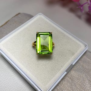 May include: A gold ring featuring a large, emerald-cut green gemstone. The ring is presented in a white box.