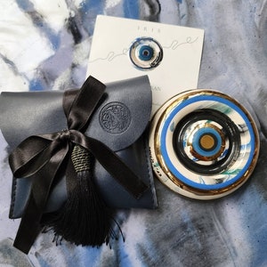 May include: A blue and white ceramic evil eye charm with gold accents, packaged in a blue leather pouch with a black tassel and a black ribbon. The pouch has a gold embossed logo that says "Iris Studio".