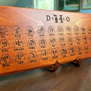 May include: Wooden Diablo II rune board with engraved runes and the game's logo. The board is stained a reddish-brown color and is supported by a brown stand. The runes are etched into the wood.