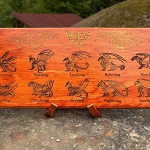 May include: A rectangular wooden plaque featuring the laser-engraved text Wings of Fire and ten distinct dragon illustrations. Each dragon is labeled with its tribe name: Hivewing, Silkwing, Leafwing, Rainwing, Seawing, Icewing, Mudwing, Skywing, Nightwing, and Sandwing.