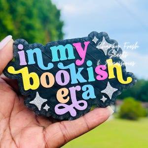 May include: A black, cloud-shaped plaque featuring the colorful, raised text "in my bookish era" in shades of purple, blue, yellow, and pink. The design is accented with silver glitter star shapes and includes the text "Chunky Fresh Scents & Accessories".