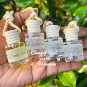 Peut inclure: Quatre petits flacons diffuseurs de parfum pour voiture en verre avec bouchons en bois et cordons de suspension. Chaque flacon porte une étiquette avec le texte Chunky Fresh Scents et les noms de parfums : Strawberry Poundcake, Watermelon Lemonade, Fifty Shades et Mahogany Teakwood.