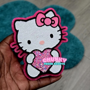 Puede incluir: Un objeto brillante blanco y rosa con forma de Hello Kitty, con un lazo rosa y un corazón. El objeto tiene contornos negros y el texto "CHUNKY FRESH SCENTS" en rosa y azul.