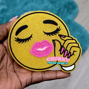 May include: A yellow emoji-shaped air freshener with closed eyes, long eyelashes, and pink glitter lips. The emoji is held in a hand and has the text "CHUNKY FRESH SCENTS" in a white and teal box.