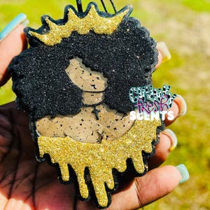 May include: A decorative air freshener shaped like a silhouette of a person with a large afro hairstyle, wearing a gold glitter crown and a cross necklace. The design features black glitter, tan cork-like texture, and gold glitter drips.