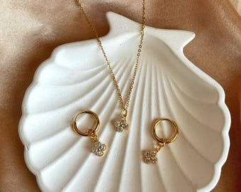 May include: A gold-toned jewelry set featuring a necklace and two hoop earrings, each adorned with a four-leaf clover pendant embellished with small, sparkling stones. The jewelry rests on a white, seashell-shaped dish.