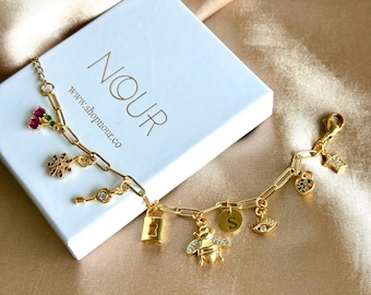 May include: A gold-tone chain bracelet featuring multiple charms including a bee, a padlock, a letter S disc, an eye, a key, a flower, cherries, and a star, resting on a white box labeled NOUR with the text www.shopnour.co.