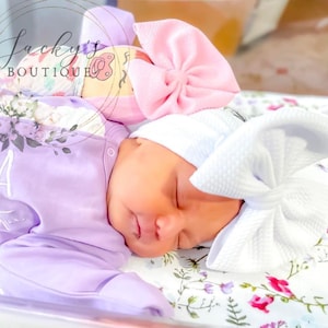 May include: A lavender long-sleeve infant sleeper with white ruffles and the embroidered name Rosalia Violet. The infant wears a white textured beanie with a large matching bow. A pink bow is also visible near the infant's head.